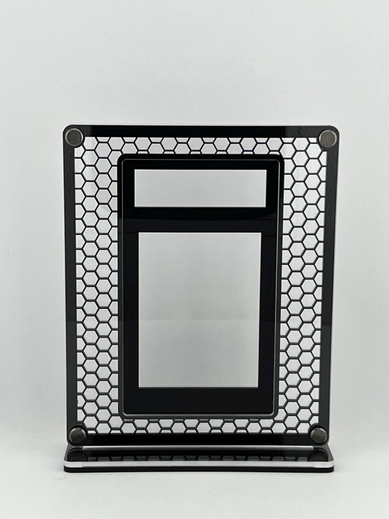Puede incluir: Un marco de fotos de metal negro con un dise&ntilde;o de malla de panal. El marco tiene forma rectangular con una abertura rectangular m&aacute;s peque&ntilde;a en el interior. El marco est&aacute; soportado por una base con una franja blanca.