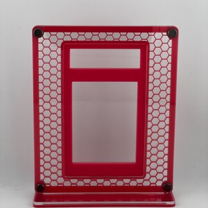 Puede incluir: Un marco de fotos rojo brillante con un dise&ntilde;o &uacute;nico de panal de abeja. El marco es rectangular con un marco interior m&aacute;s peque&ntilde;o y una base. Se ven peque&ntilde;os imanes negros en las esquinas. El marco mide unos 15 cm de alto.