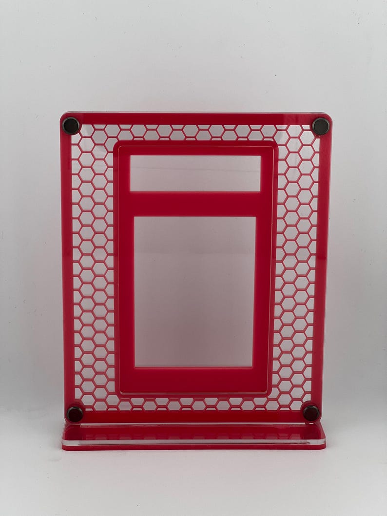 Puede incluir: Un marco de fotos rojo brillante con un dise&ntilde;o &uacute;nico de panal de abeja. El marco es rectangular con un marco interior m&aacute;s peque&ntilde;o y una base. Se ven peque&ntilde;os imanes negros en las esquinas. El marco mide unos 15 cm de alto.