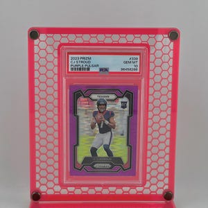 Puede incluir: Un marco de exhibici&oacute;n rosa que contiene una tarjeta coleccionable 2023 Prizm CJ Stroud Purple Pulsar. La tarjeta presenta una foto de un jugador de f&uacute;tbol americano con el uniforme de los Texans. El marco tiene un dise&ntilde;o de panal.