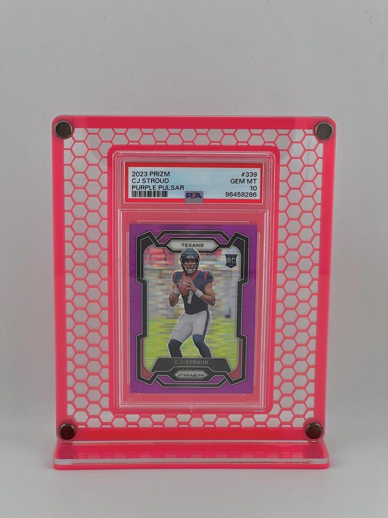 Puede incluir: Un marco de exhibici&oacute;n rosa que contiene una tarjeta coleccionable 2023 Prizm CJ Stroud Purple Pulsar. La tarjeta presenta una foto de un jugador de f&uacute;tbol americano con el uniforme de los Texans. El marco tiene un dise&ntilde;o de panal.