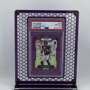 Puede incluir: Un marco de exhibici&oacute;n morado con un dise&ntilde;o de panal, que contiene una tarjeta coleccionable deportiva. La tarjeta presenta a un jugador de f&uacute;tbol americano con el uniforme de los Texans. La tarjeta dice "2023 Prizm CJ Stroud Purple Pulsar".