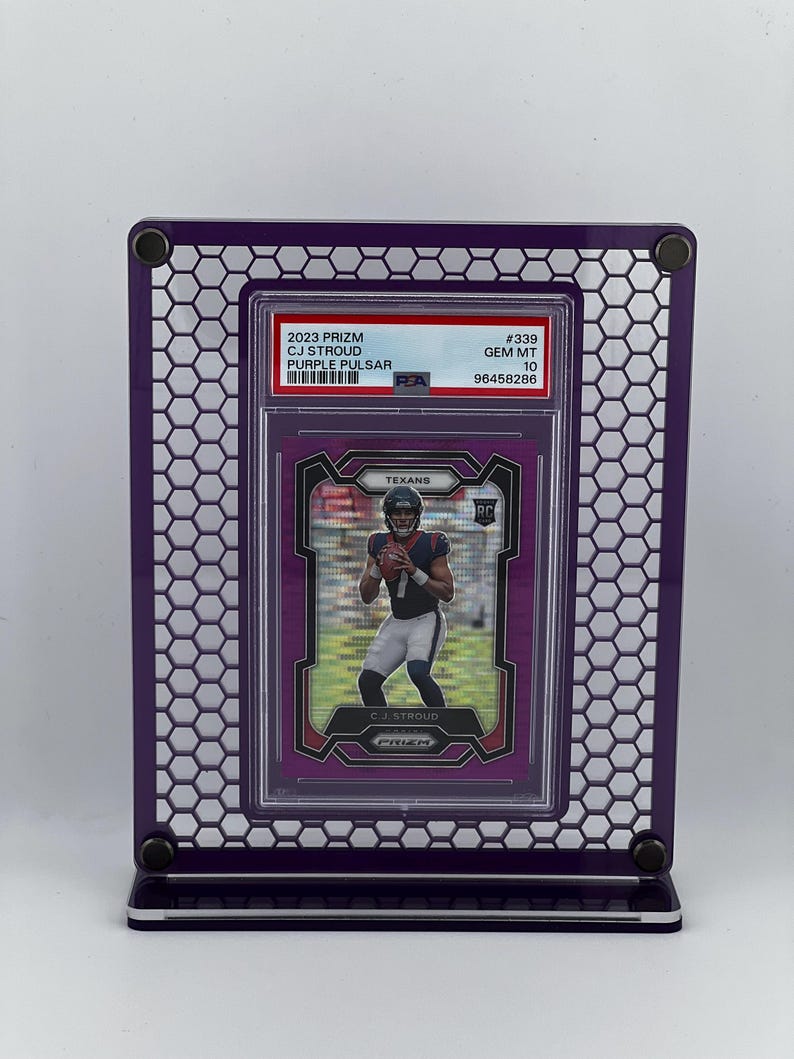 Puede incluir: Un marco de exhibici&oacute;n morado con un dise&ntilde;o de panal, que contiene una tarjeta coleccionable deportiva. La tarjeta presenta a un jugador de f&uacute;tbol americano con el uniforme de los Texans. La tarjeta dice "2023 Prizm CJ Stroud Purple Pulsar".