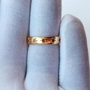 May include: A gold ring featuring small, red gemstones set into the band. The ring has a repeating starburst pattern. The ring is shown on a white background.