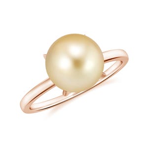 Puede incluir: Un anillo de oro rosa con una perla dorada grande y redonda. La perla es el punto focal, engastada en una banda delicada con un diseño simple y elegante. El anillo se muestra sobre un fondo blanco.