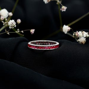 May include: A silver eternity ring featuring a continuous line of bright red gemstones. The ring is displayed against a dark backdrop, with small white flowers in the background. The gemstones are uniformly set around the ring's circumference.