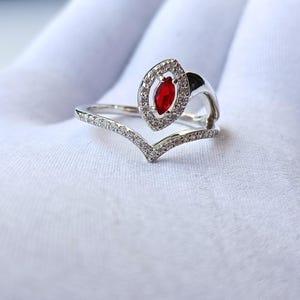 May include: A silver ring with a marquise-shaped red gemstone at the center, surrounded by a halo of small, clear stones. The band features a V-shaped design with additional clear stones. The ring is set against a white background.
