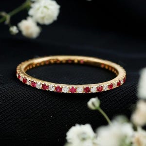 Op de afbeelding: Een gouden bandring met afwisselend rode en witte edelstenen. De ring staat tegen een zwarte achtergrond, met witte bloemen op de achtergrond. De edelstenen zijn gelijkmatig verdeeld over de band.