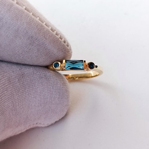 May include: A gold ring featuring a rectangular blue gemstone flanked by two smaller, round blue gemstones. The ring has a delicate band and is set against a white background.