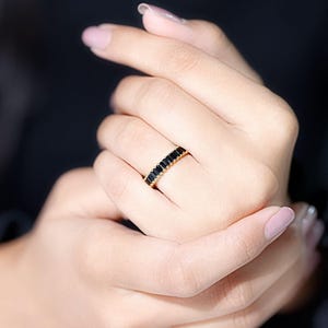 以下が含まれることがあります： 黒い長方形の石が並んだゴールドリング。暗い背景の中で、人の指にはめられています。シンプルでエレガントなデザインで、普段使いにも特別な場面にも適しています。