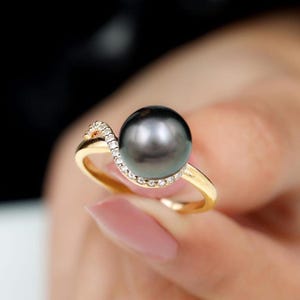 Könnte beinhalten: Ein Goldring mit einer großen, runden, dunkelgrauen Perle als Herzstück. Der Ring besteht aus poliertem Gelbgold und verfügt über ein geschwungenes Element, das mit kleinen, klaren, funkelnden Steinen besetzt ist und die Perle einrahmt.