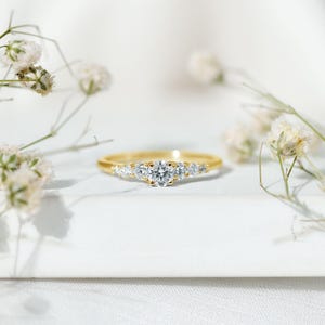 May include: A gold ring with a central round diamond and smaller diamonds on either side. The ring is set against a white background with delicate white flowers, creating a soft, elegant aesthetic. The ring is a classic design.