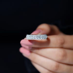 May include: A close-up of a diamond eternity ring. The ring features multiple rows of small, sparkling diamonds set in a silver band. The ring is held against a dark background, highlighting its brilliance.