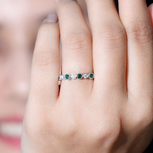 Può includere: Un anello d'argento con una serie di pietre preziose verdi e chiare alternate. L'anello è incastonato con piccole pietre rotonde. Le pietre preziose sono distanziate uniformemente attorno alla fascia. L'anello è sul dito di una persona.