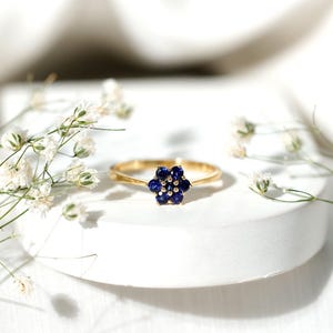 Op de afbeelding: Een gouden ring met een bloemvormige cluster van donkerblauwe edelstenen. De ring ligt op een wit oppervlak, met kleine witte bloemen op de achtergrond.