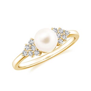 Op de afbeelding: Een gouden ring met een grote, ronde witte parel als middelpunt. Aan weerszijden van de parel bevinden zich clusters van kleine, heldere edelstenen. De ring heeft een gladde, gepolijste band.