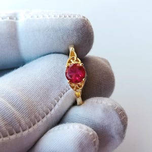 May include: A gold-colored ring featuring a round, vibrant red gemstone. The ring has an intricate Celtic knot design on the band and around the gemstone setting. The ring is held by a gloved hand.