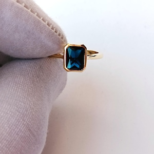 Può includere: Un anello d'oro con una pietra preziosa rettangolare blu scuro. La pietra è incastonata in una lunetta d'oro e la fascia dell'anello è in oro lucido. L'anello è mostrato su sfondo bianco.