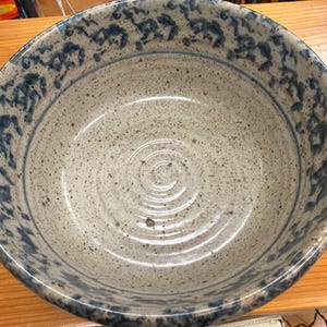 May include: A large, speckled stoneware bowl with a wide rim. The bowl's interior is a light beige with dark brown speckles. The rim features a repeating pattern of dark blue floral designs. The bowl is sitting on a wooden surface.