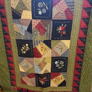 May include: A handcrafted quilt with a patchwork design. The quilt features squares of various fabrics in shades of green, red, blue, and tan. Each square has embroidered floral details. The border is a dark green with red triangle accents.