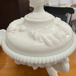 May include: A white, antique, covered serving dish with a decorative lid and base. The lid features a finial and embossed floral and leaf designs. The dish rests on three feet and has a beaded edge. A classic piece for serving or display.
