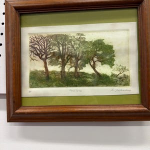 May include: Framed artwork featuring a landscape with trees in various stages of foliage. The artwork is set against a green mat and enclosed in a dark brown wooden frame. The text "Four Trees" is visible.