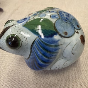 May include: A ceramic frog figurine with a white base, adorned with blue floral patterns and green leaves. The frog has large, dark eyes and is painted with intricate details. The figurine is a decorative item.