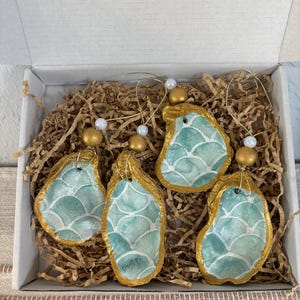 Könnte beinhalten: Vier Austernschalen-Ornamente mit goldenem Rand und einem türkisfarbenen Schuppenmuster, präsentiert in einer weißen Box mit braunem Papierfüllmaterial. Jedes Ornament hat eine goldene Perle und eine weiße Perle.