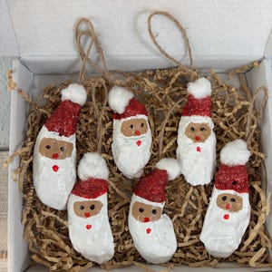 Könnte beinhalten: Sechs handgefertigte Weihnachtsschmuckstücke in Form von Weihnachtsmännern. Jedes Ornament hat einen weißen Körper, einen roten Glitzerhut und einen kleinen weißen Bommel. Die Gesichter sind mit braunen, schwarzen und roten Details bemalt. Präsentiert in einer weißen Box.
