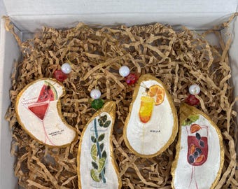 May include: A set of four hand-painted oyster shell ornaments, each depicting a different cocktail: watermelon, mojito, mimosa, and jamaica. The shells have gold edges and are displayed in a box with brown paper filler. Each ornament has a beaded string for hanging.
