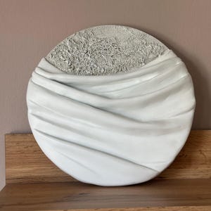 Può includere: Un'opera d'arte murale circolare caratterizzata da una sezione superiore grigio chiaro testurizzata con una superficie ruvida e craterizzata, e una sezione inferiore bianca liscia con pieghe profonde e fluide che imitano il tessuto drappeggiato.