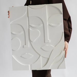 Puede incluir: Una pieza de arte mural rectangular de color blanco que presenta una escultura en relieve de dos rostros abstractos. El diseño consiste en líneas y formas suaves y elevadas que forman rasgos faciales, incluyendo ojos cerrados, narices y labios.
