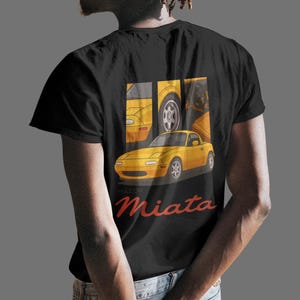 May include: Black t-shirt featuring a yellow Miata sports car graphic. The design includes a full car image and car part details. The word "Miata" is written in red script below the car image. The shirt is made of a soft material.