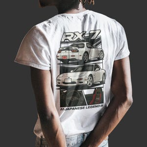 May include: White t-shirt featuring a graphic of two white sports cars with the text "RX-7" and "JAPANESE LEGEND II". The cars are displayed on a black and white background. The shirt is made of a soft material.
