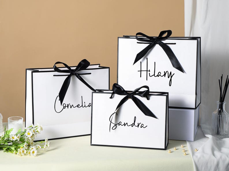 May include: Three white gift bags with black ribbon bows and black trim. The bags are personalised with the names "Hilary", "Cornelia", and "Sandra" in black script.