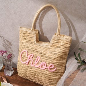 May include: A beige woven straw tote bag featuring two top handles and the name Chloe embroidered in pink cursive script on the front. The bag has an open top design with a structured rectangular shape and a natural tan fiber texture throughout.