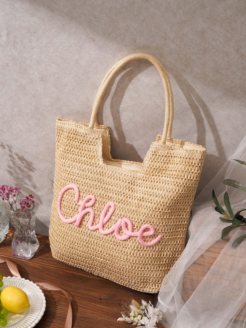 May include: A beige woven straw tote bag featuring two top handles and the name Chloe embroidered in pink cursive script on the front. The bag has an open top design with a structured rectangular shape and a natural tan fiber texture throughout.