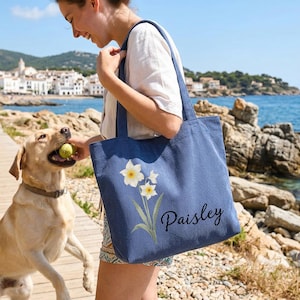 Puede incluir: Un bolso tote de pana azul con un dise&ntilde;o floral y el nombre "Paisley" bordado. La persona sostiene el bolso cerca de un perro y el mar. El perro sostiene una pelota de tenis. Al fondo, un pueblo costero.