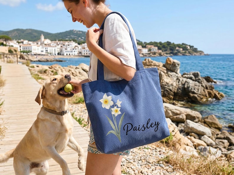 Puede incluir: Un bolso tote de pana azul con un dise&ntilde;o floral y el nombre "Paisley" bordado. La persona sostiene el bolso cerca de un perro y el mar. El perro sostiene una pelota de tenis. Al fondo, un pueblo costero.