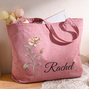 Puede incluir: Un bolso tote de pana rosa con un dise&ntilde;o floral y el nombre "Rachel" en cursiva. La bolsa contiene un libro y tulipanes. Un peque&ntilde;o calendario de madera muestra "ABR".