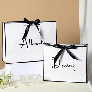 May include: Two white gift bags with black ribbon bows and black trim. The larger bag has the text "Alberta" printed on it. The smaller bag has the text "Destiny" printed on it.