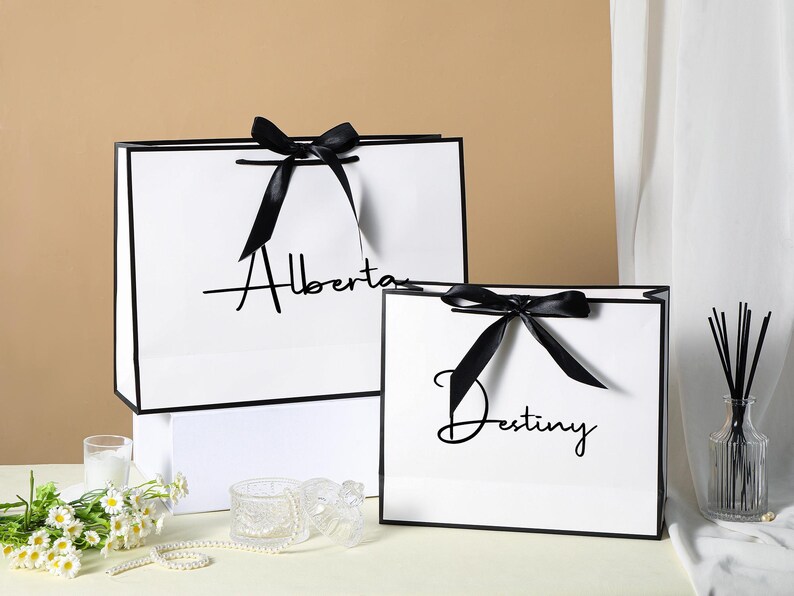 May include: Two white gift bags with black ribbon bows and black trim. The larger bag has the text "Alberta" printed on it. The smaller bag has the text "Destiny" printed on it.