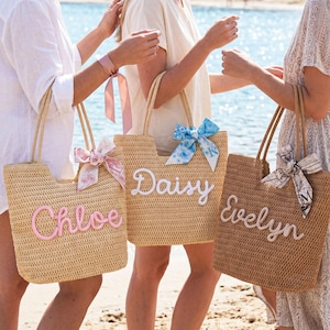 May include: Three woven straw tote bags with rope handles. The left bag is light tan with pink script text reading Chloe and a pink ribbon. The center bag is tan with white script text reading Daisy and a blue floral ribbon. The right bag is brown with white script text reading Evelyn and a patterned ribbon.