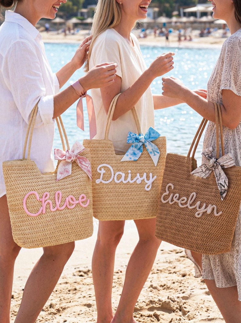 May include: Three woven straw tote bags with rope handles. The left bag is light tan with pink script text reading Chloe and a pink ribbon. The center bag is tan with white script text reading Daisy and a blue floral ribbon. The right bag is brown with white script text reading Evelyn and a patterned ribbon.