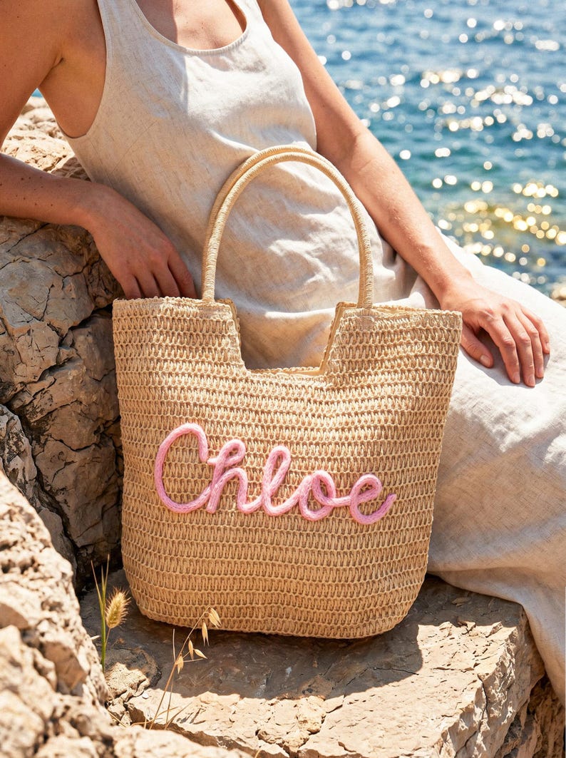 May include: A beige woven straw tote bag featuring two top handles and the name Chloe written in raised pink cursive script on the front. The bag has a structured rectangular shape with an open top and a textured, natural fiber construction.