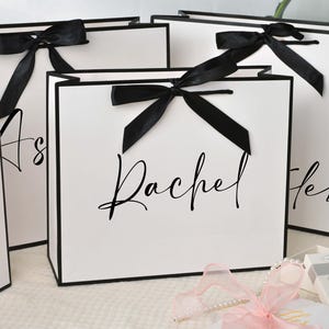 May include: White gift bags with black trim and black satin bows. The bags have names written in elegant script. A pink ribbon and small gift boxes are in the foreground.