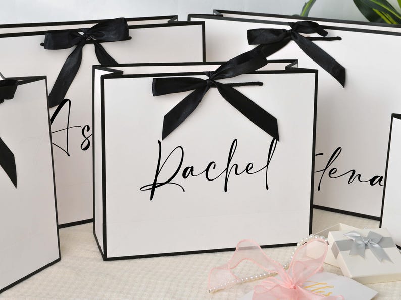 May include: White gift bags with black trim and black satin bows. The bags have names written in elegant script. A pink ribbon and small gift boxes are in the foreground.