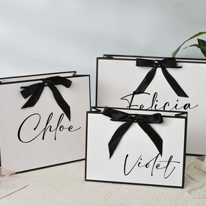 May include: Three white gift bags with black trim and black bows. The bags are labeled with the names Chloe, Felicia, and Violet in elegant script. A small gift box and a string of pearls are also visible.