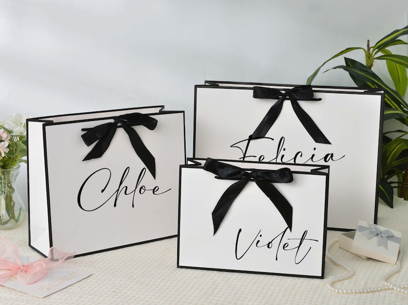 May include: Three white gift bags with black trim and black bows. The bags are labeled with the names Chloe, Felicia, and Violet in elegant script. A small gift box and a string of pearls are also visible.
