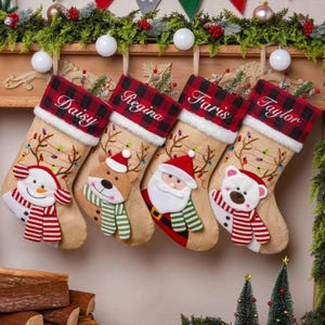 Op de afbeelding: Vier kerstsokken van jute met rood-zwart geruite boorden, witte imitatiebontranden en de geborduurde namen Daisy, Regina, Faris en Taylor. Elke sok is voorzien van een pluche figuur zoals een sneeuwpop, rendier, kerstman of ijsbeer.